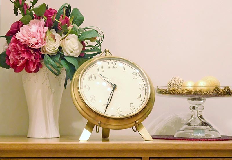 A decorative tabletop display is arranged on a light wood surface against a plain, off-white wall. In the center sits a large, vintage-style round clock with a brass-colored metal frame and legs.