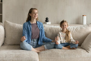 Air Conditioning Noise Concerns - Relaxed peaceful young mother and little cute child daughter sitting in lotus position on cozy couch with closed eyes making mudra gesture, doing yoga breathing exercises, enjoying mindful moment.