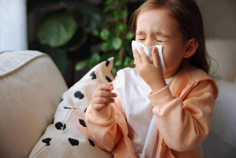 A young girl sitting on a couch, squeezing her eyes shut as she blows or wipes her nose with a tissue.
