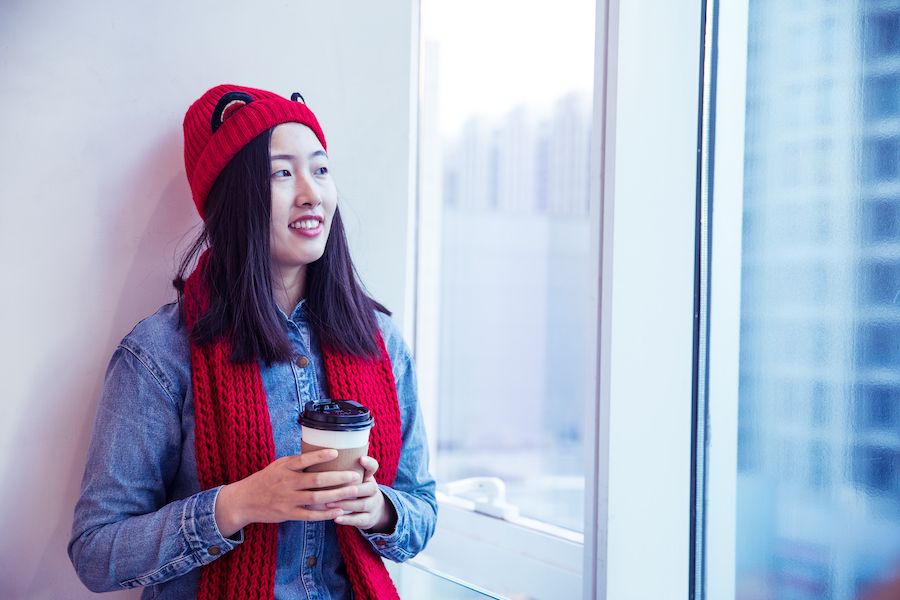 The Basics of Winter Indoor Air Quality. Image of woman inside looking out window while wearing a hat and scarf and holding a coffee.